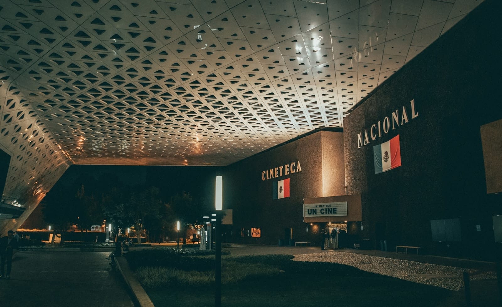 Vista notturna della Cineteca Nacional a Città del Messico, con il suo moderno tetto geometrico, l'ingresso illuminato e le bandiere messicane sopra l'edificio principale.