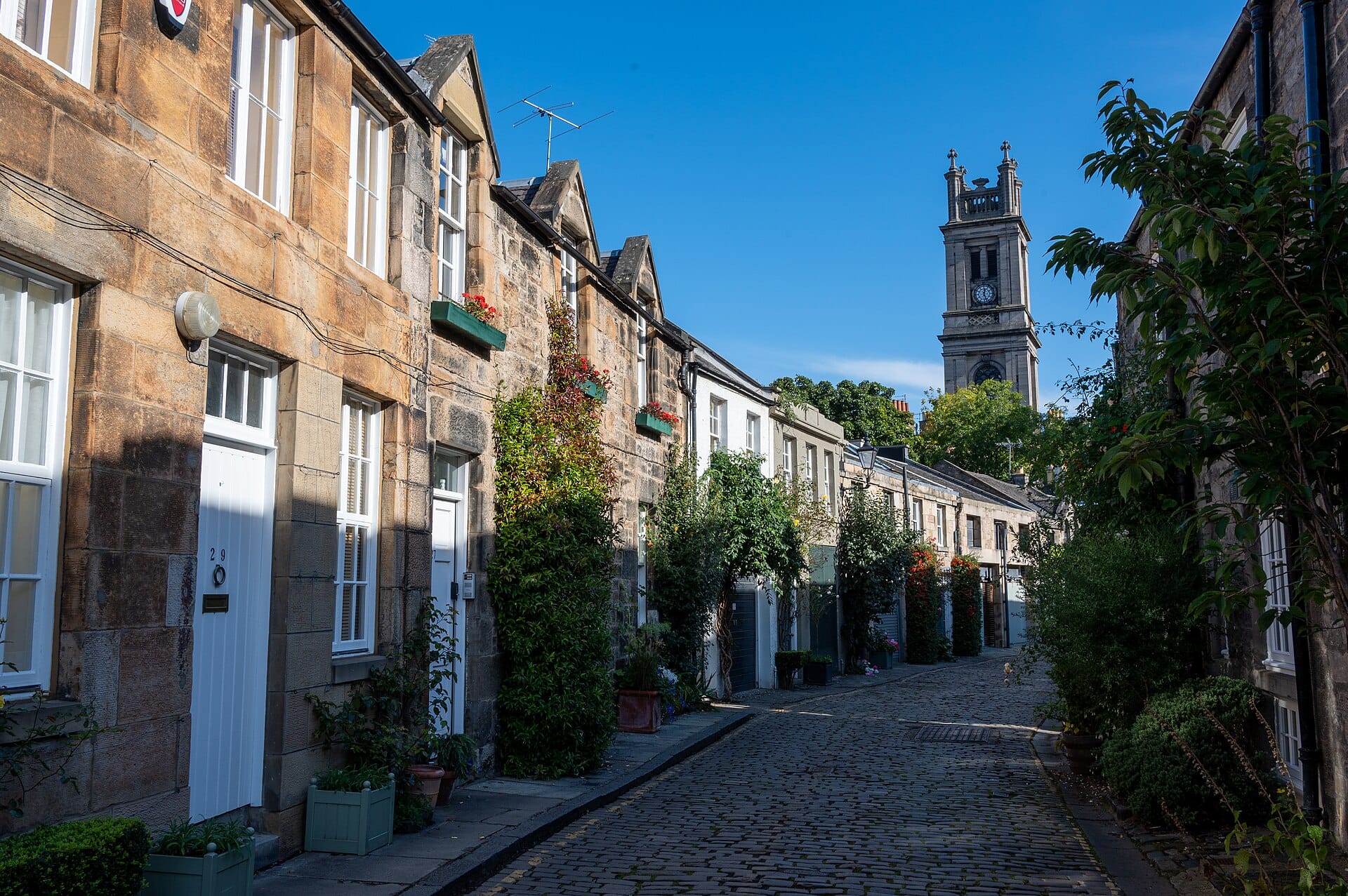 Circus Lane in Edinburgh an einem sonnigen Tag: das Kopfsteinpflaster, gesäumt von Steinhäuschen, Blumenkästen, Grünpflanzen und dem Turm der St Stephen's Church im Hintergrund.