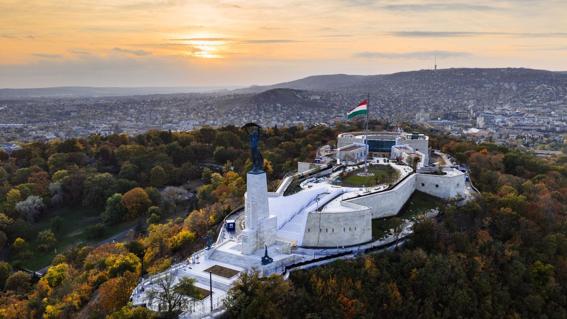 Vista aerea della fortezza della Citadella e della Statua della Libertà in cima alla collina di Gellért a Budapest al tramonto, con alberi dai colori autunnali vivaci e il panorama urbano sullo sfondo.