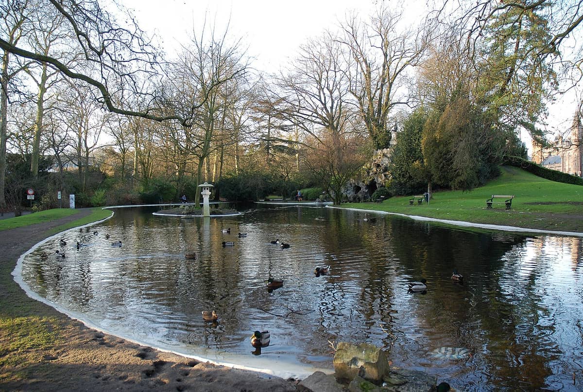 Citadelpark pond in Ghent with swimming ducks, green lawns, benches, leafless trees, and a small stone structure in soft daylight.