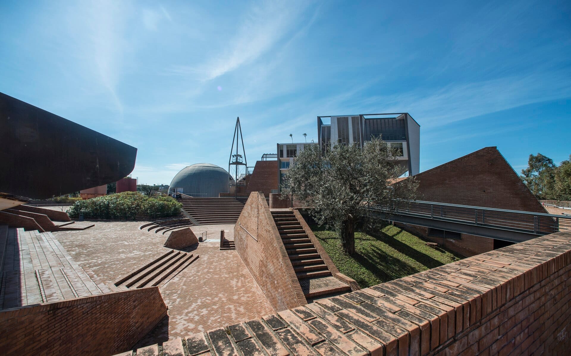 L'esterno moderno della Città della Scienza a Napoli con vialetti in mattoni, gradinate ad anfiteatro, verde e l'iconica cupola del planetario in una giornata di sole.