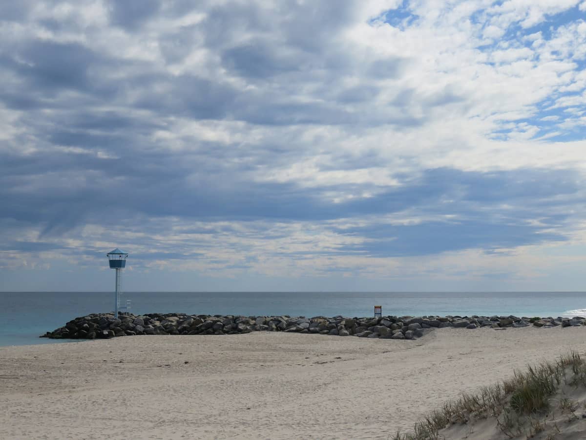 Breiter Sandstrand an der City Beach, Perth, mit einem felsigen Wellenbrecher, einem Rettungsturm und bewölktem Himmel über dem Indischen Ozean.