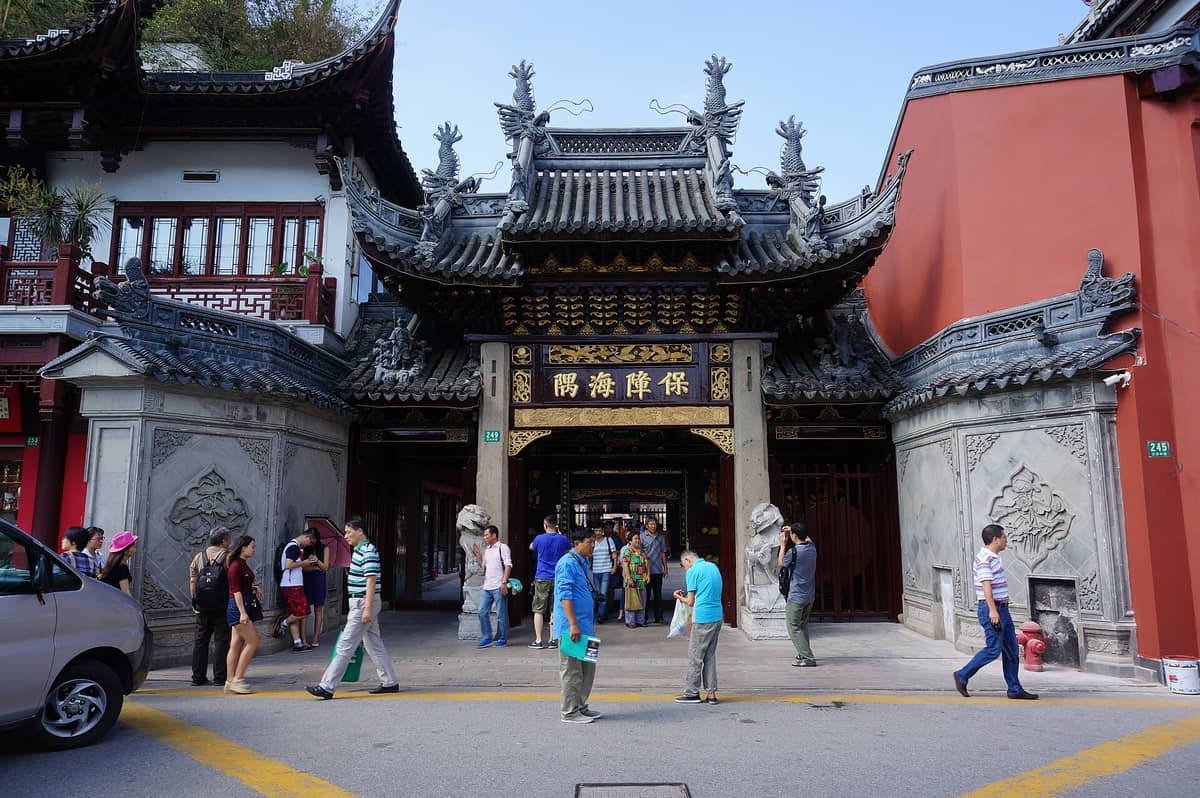 Haupteingangstor des Stadtgotttempels Shanghai mit kunstvoll verziertem traditionellen Dach, Drachenskulpturen, goldenen chinesischen Schriftzeichen und Besuchern, die den Tempelkomplex betreten.