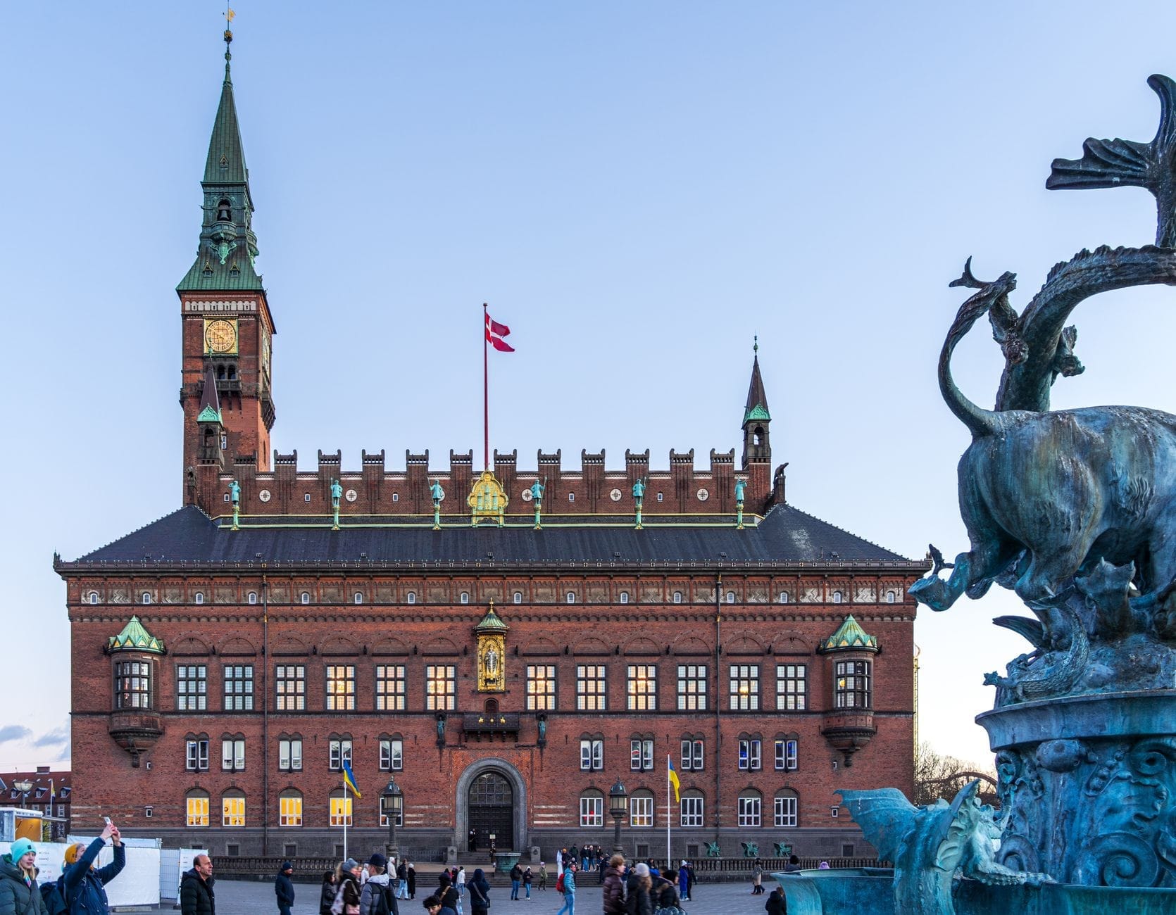 Il Municipio di Copenaghen visto da Rådhuspladsen, con persone che camminano nella piazza e la fontana del drago in primo piano.