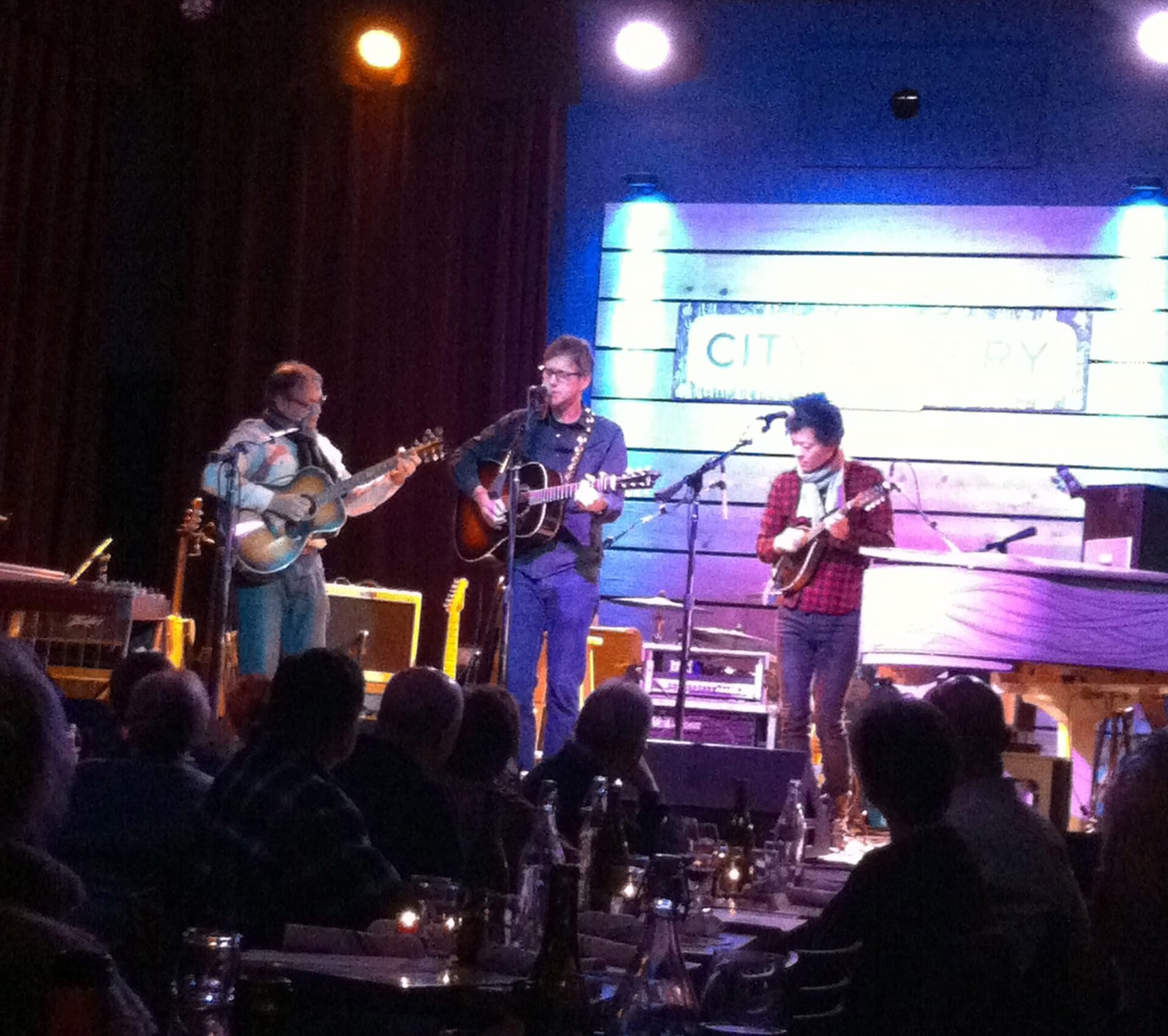 Tres músicos tocan con guitarras y mandolina en el escenario cálidamente iluminado de City Winery Nashville, mientras el público se sienta en mesas con velas.