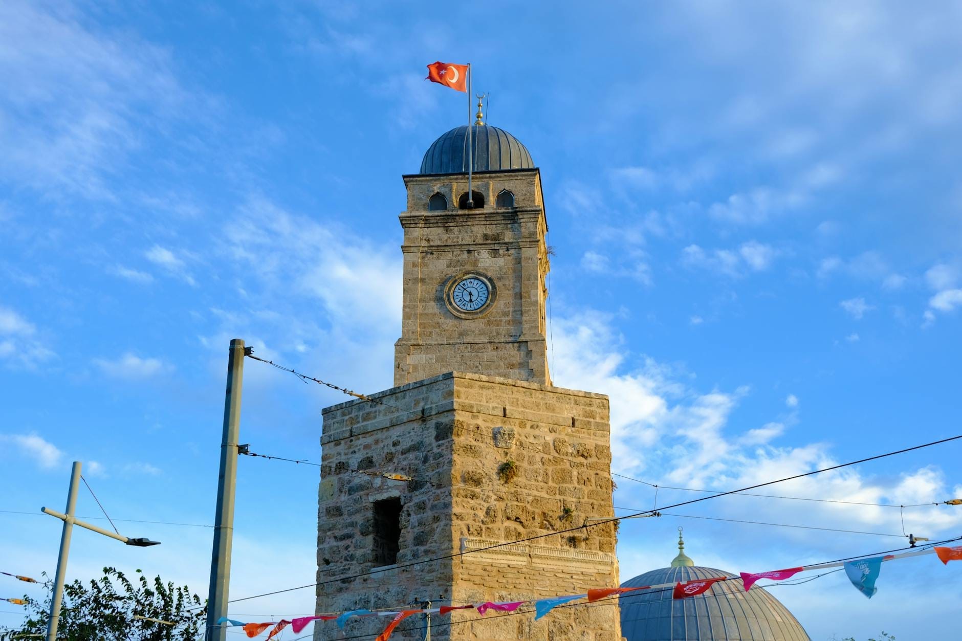 La torre dell'orologio ottomana Saat Kulesi ad Antalya, con la bandiera turca in cima, un cielo luminoso sullo sfondo e ghirlande festose in primo piano.
