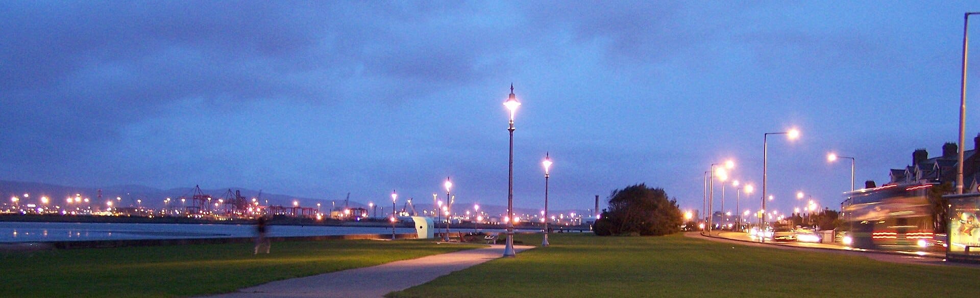 Lungomare di Clontarf al crepuscolo con lampioni illuminati, un sentiero erboso accanto al mare, luci della città e traffico sotto un cielo blu.