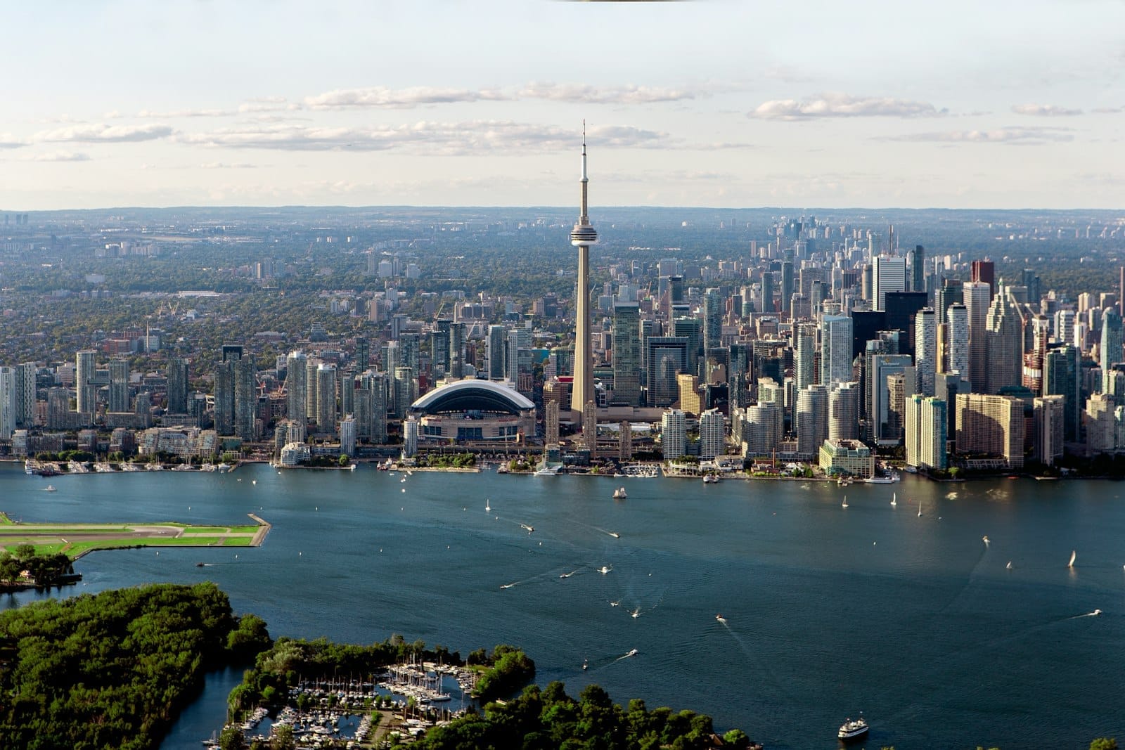 ภาพมุมสูงของโตรอนโต เห็น CN Tower เด่นเหนือเส้นขอบฟ้า เมือง ทะเลสาบออนตาริโอ และเรือในวันที่ฟ้าใส