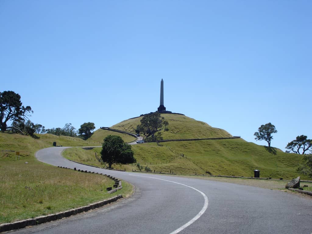 One Tree Hill / Maungakiekie am Coast to Coast Walkway in Auckland: ein grasbewachsener Vulkankegel mit Obelisk auf dem Gipfel, einer gewundenen asphaltierten Straße und vereinzelten Bäumen unter klarem blauem Himmel.