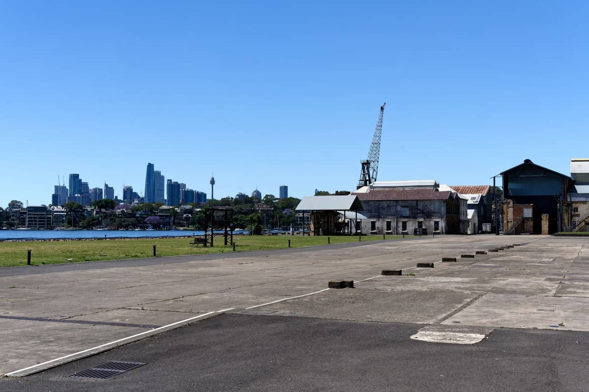 Vista panorámica de la isla Cockatoo con edificios industriales patrimoniales, una grúa en el muelle y el skyline de Sídney al fondo bajo un cielo azul.