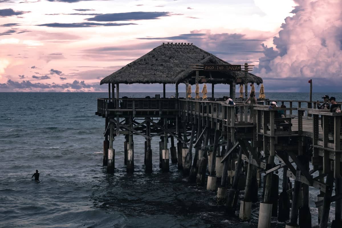Cocoa Beach Pier extending out over the Atlantic Ocean at sunset, with people on the pier and a surfer in the water below.