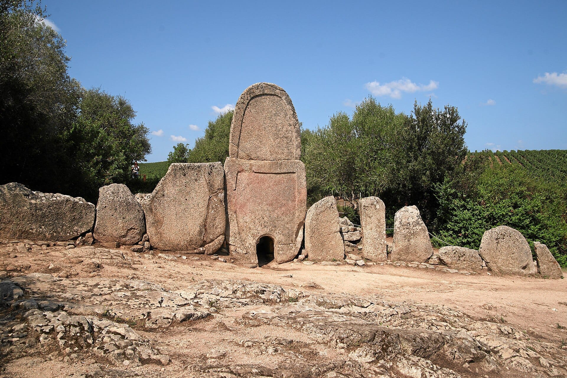 Vorderansicht des Riesengrabes Coddu Vecchiu mit großen Granitsteinen, die in einem Halbkreis zwischen grünen Bäumen und blauem Himmel stehen.