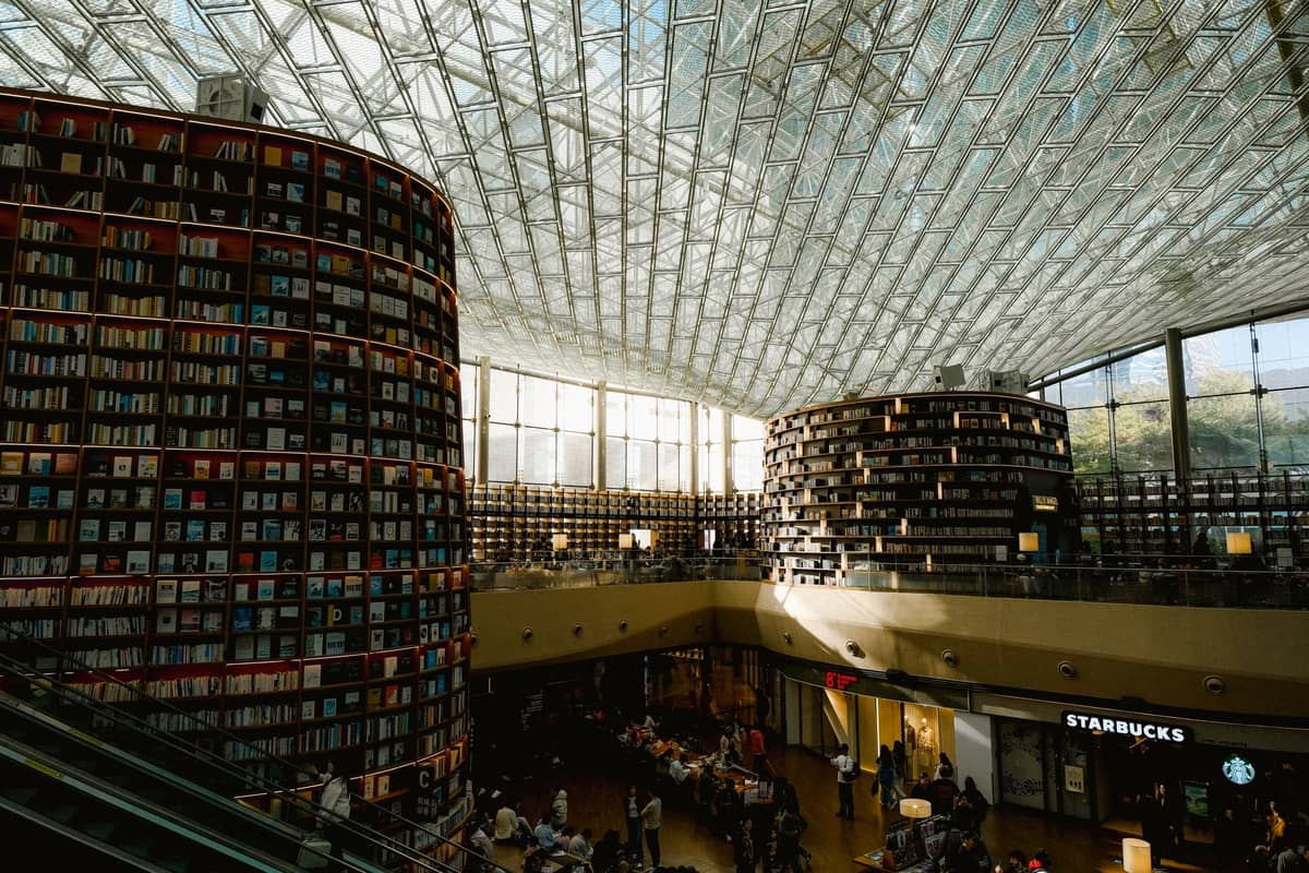 Vista ampla da Biblioteca Starfield no COEX Mall, com duas enormes estantes circulares se erguendo em direção ao teto de vidro e uma movimentada área de compras abaixo.
