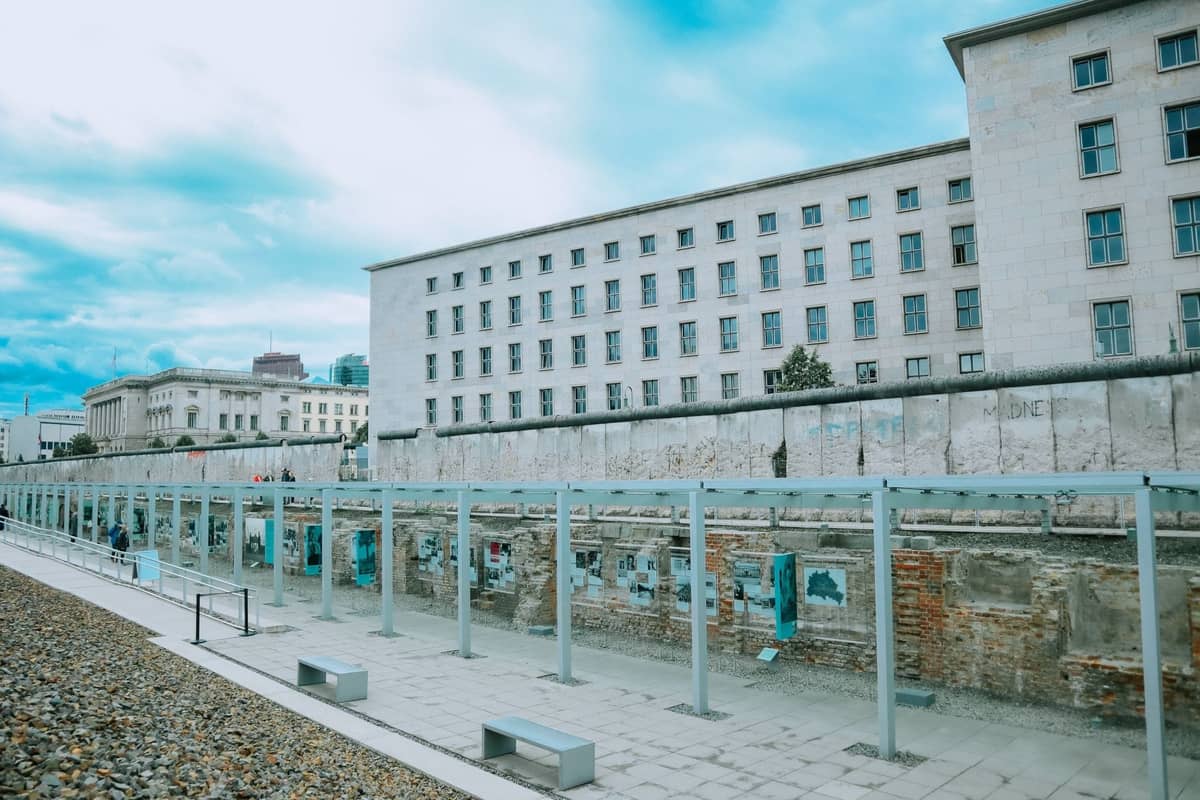Verbleibender Abschnitt der Berliner Mauer mit historischen Ausstellungen und Stadtgebäuden im Hintergrund unter einem bewölkten blauen Himmel.