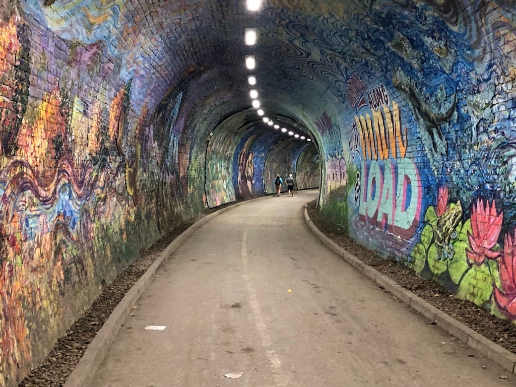 Bunte Wandgemälde bedecken die geschwungenen Backsteinwände und die Decke des Colinton Tunnel, mit einem gepflasterten Weg und Besuchern, die im Inneren unter hellem Licht spazieren.
