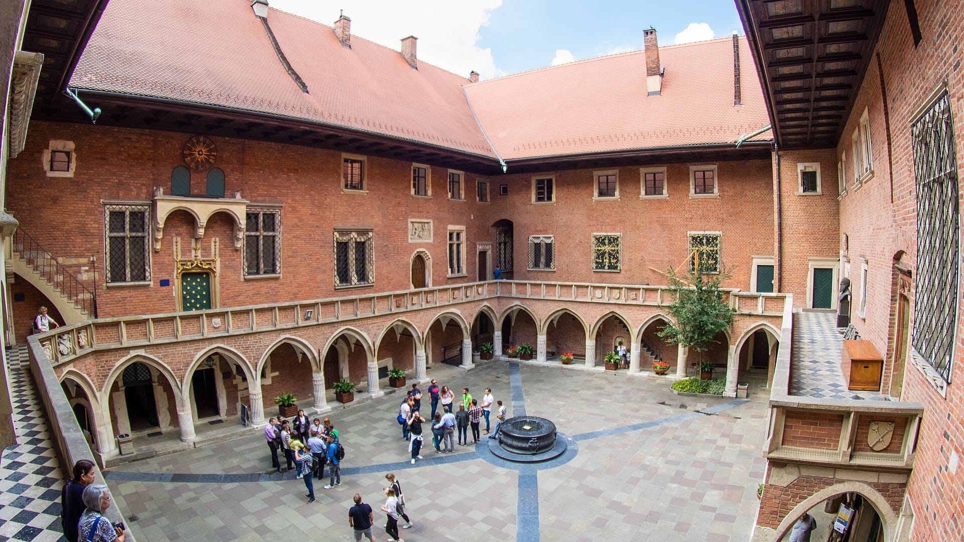 ลาน Collegium Maius พร้อมซุ้มอิฐแดงแบบโกธิก แกลเลอรีโค้ง น้ำพุกลางลาน และกลุ่มนักท่องเที่ยวที่กำลังสำรวจภายในอาคารมหาวิทยาลัยประวัติศาสตร์