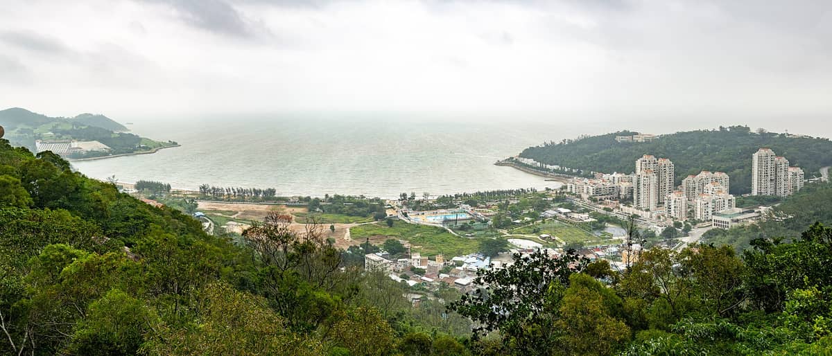 Panoramablick auf Coloane, Macau: üppige bewaldete Hügel, Wohngebäude, Sandstrände und die Küste an einem bewölkten Tag.