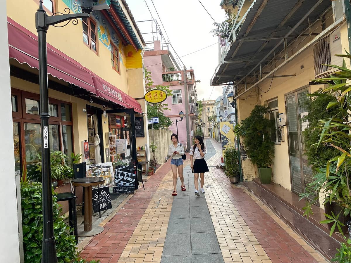 Zwei junge Frauen spazieren durch eine farbenfrohe, enge Straße, gesäumt von gelben Kolonialgebäuden und Ladengeschäften im Coloane Village, Macau.