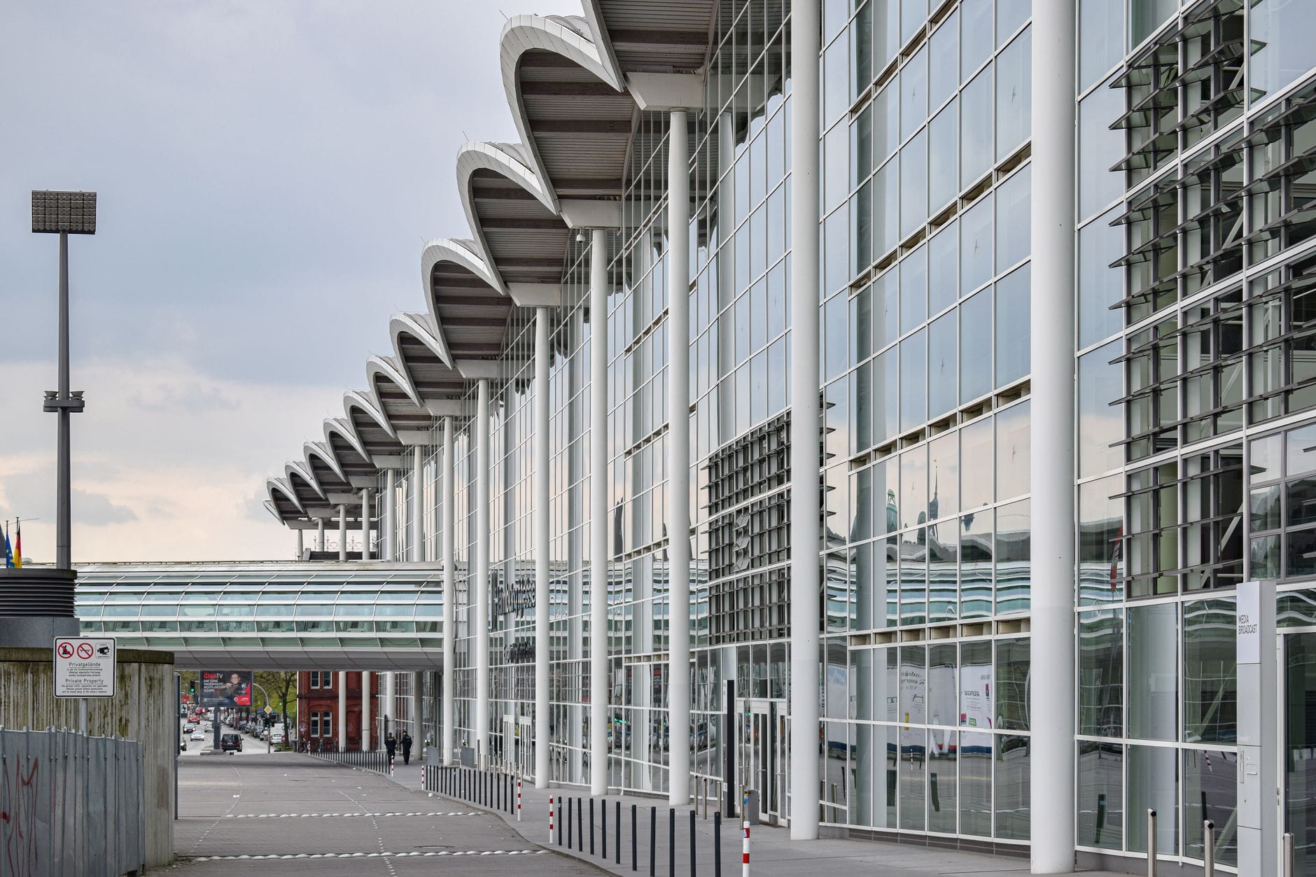 ภายนอกกระจกสมัยใหม่ของศูนย์นิทรรศการ Koelnmesse ในเมือง Cologne ประเทศเยอรมนี พร้อมทางเดินมีหลังคาและพื้นที่คนเดินเท้าที่ว่างเปล่า เน้นสิ่งอำนวยความสะดวกสำหรับงานแสดงสินค้าและกิจกรรม