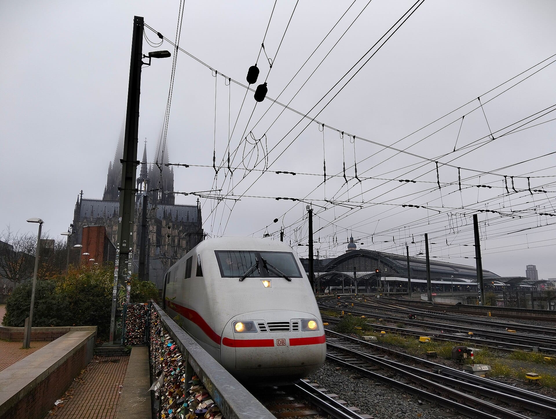 รถไฟ DB สีขาวกำลังเข้าสู่ Cologne Hauptbahnhof โดยมี Cologne Cathedral และอาคารสถานีปรากฏอยู่เบื้องหลังในวันที่มีเมฆมาก