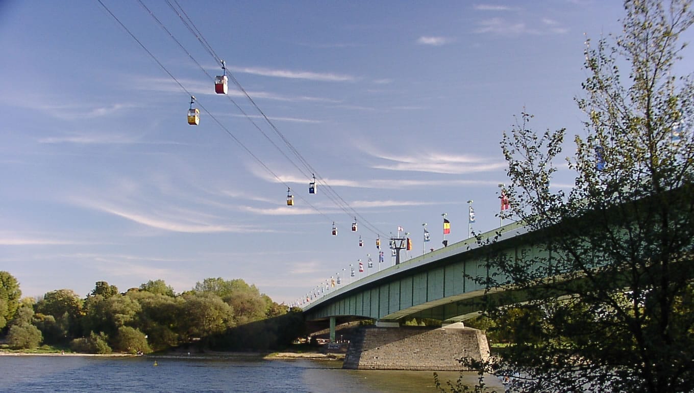 กระเช้าสีสันสดใสของ Cologne Cable Car ลอยอยู่เหนือแม่น้ำ Rhine พร้อมสะพานสีเขียว ธงชาติ และแถวต้นไม้ริมฝั่งแม่น้ำ
