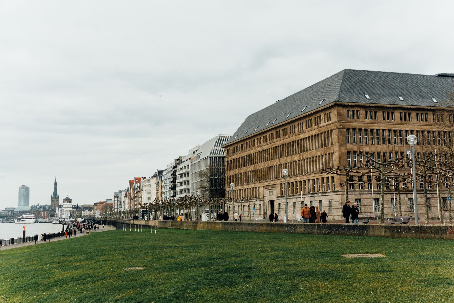 ทางเดินริมแม่น้ำที่เขียวชอุ่มในเมือง Cologne พร้อมผู้คนที่เดินอยู่บนเส้นทาง ล้อมรอบด้วยอาคารทั้งเก่าแก่และทันสมัย ริมแม่น้ำ Rhine