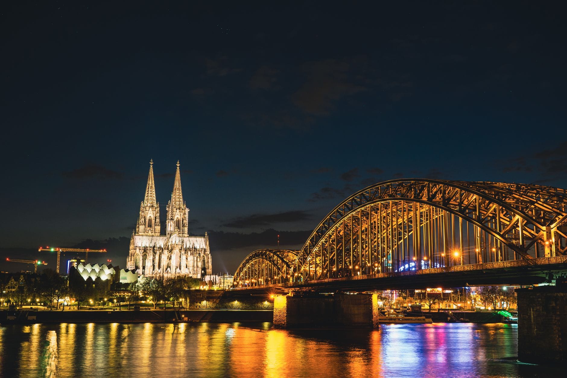 ภาพกลางคืนของมหาวิหารโคโลญและสะพาน Hohenzollern ที่สว่างไสว สะท้อนลงแม่น้ำไรน์ พร้อมแสงไฟเมืองสร้างบรรยากาศโรแมนติกในโคโลญจน์ เยอรมนี
