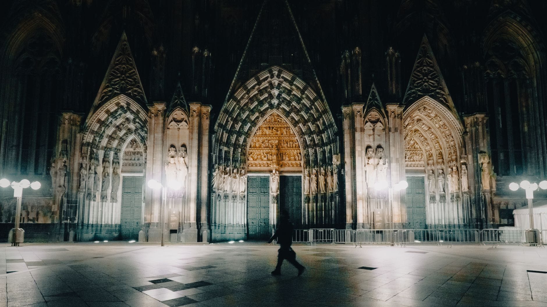 วิวยามค่ำคืนของทางเข้าหลักของ Cologne Cathedral พร้อมซุ้มโค้งแบบกอธิคอันวิจิตรและมีคนเดินผ่าน