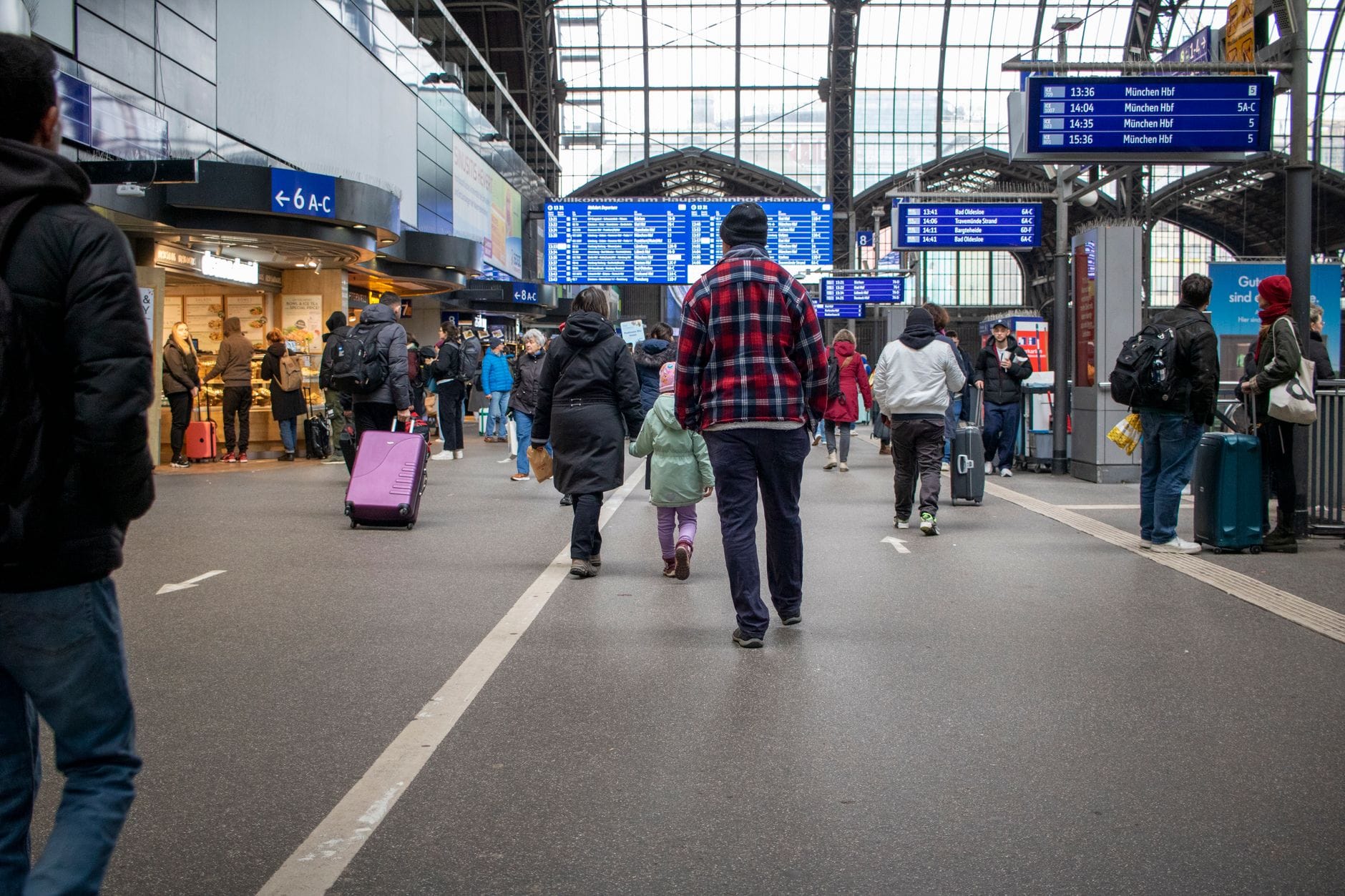 ห้องโถงภายในที่คึกคักของสถานีรถไฟขนาดใหญ่ในเยอรมนี พร้อมนักเดินทาง ป้ายข้อมูลขนาดใหญ่ และร้านค้าภายในสถานีที่มองเห็นได้
