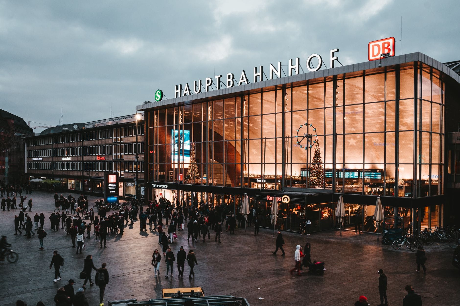 ด้านนอกของ Cologne Hauptbahnhof (Köln Hbf) ยามพลบค่ำ ด้านหน้ากระจกสว่างไสว มีผู้คนมากมายบนลานสถานีที่คึกคัก