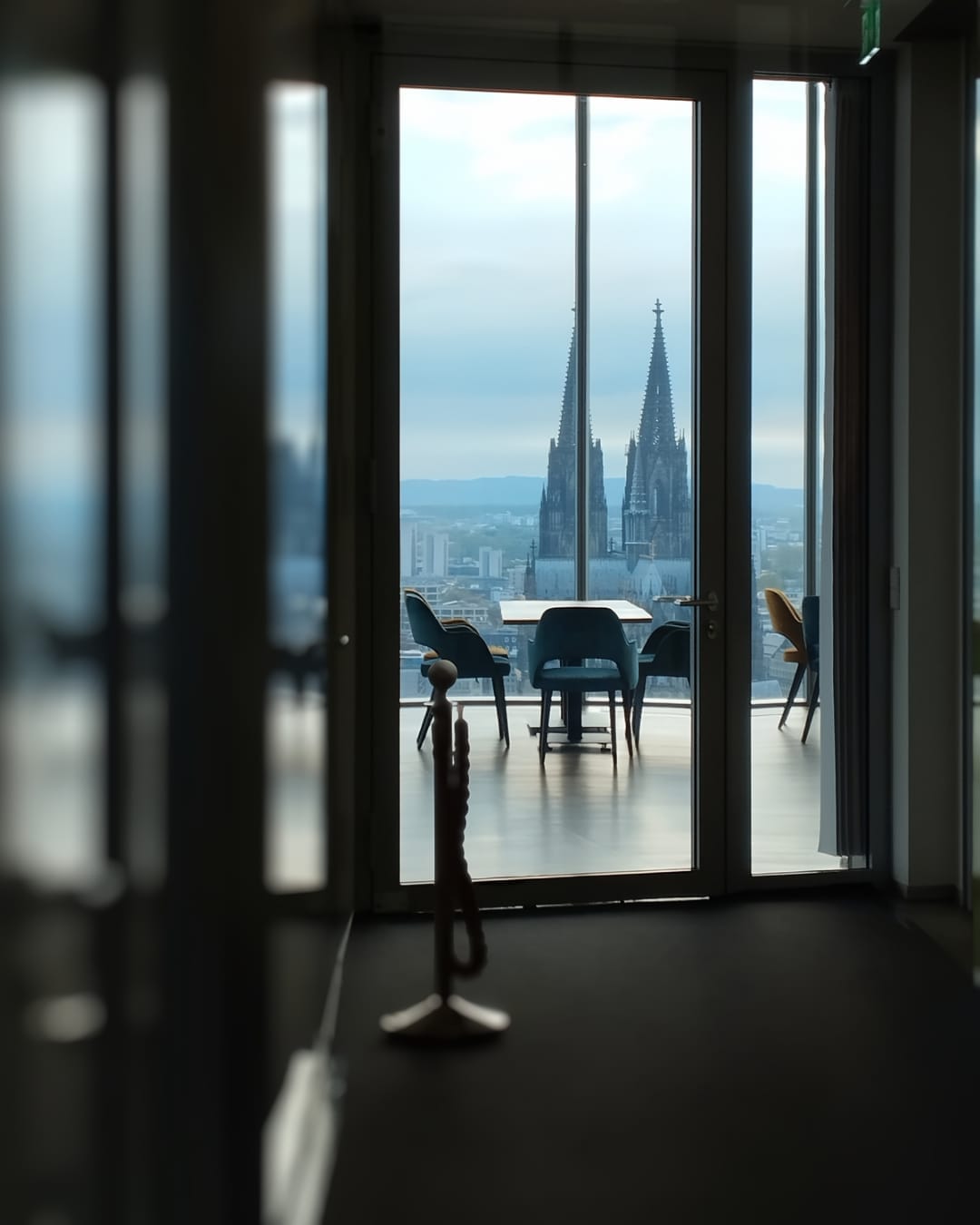 วิวผ่านประตูกระจกขนาดใหญ่ที่ Cologne Triangle Panorama แสดงให้เห็นเก้าอี้สมัยใหม่และแนวสายตาที่ชัดเจนไปยังยอดแฝดของ Cologne Cathedral