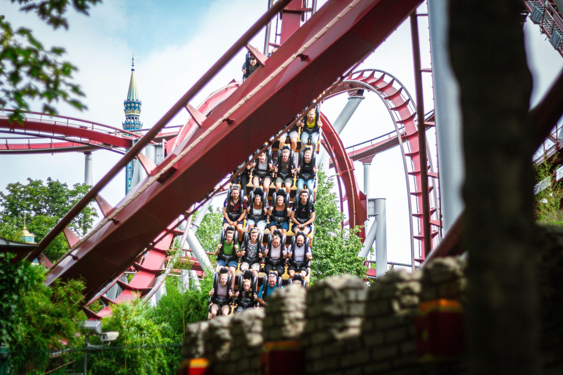 รถไฟเหาะสีแดงที่ Phantasialand พร้อมผู้โดยสารจำนวนมากกำลังลงจากส่วนที่ชันล้อมรอบด้วยต้นไม้และองค์ประกอบตกแต่งตามธีม