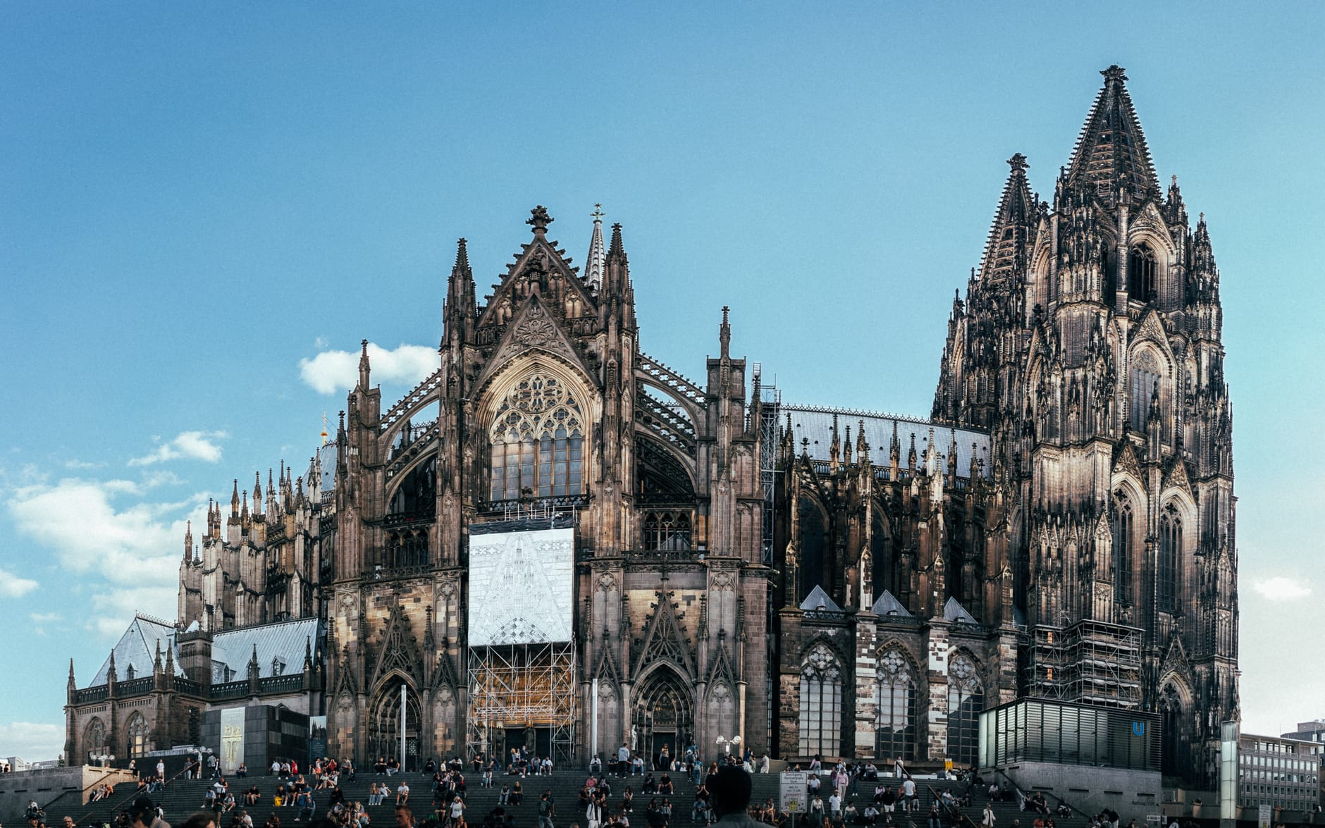 มุมมองกว้างของภายนอก Cologne Cathedral ที่มียอดแหลมสูงสองยอด จัตุรัสคึกคัก และท้องฟ้าสีฟ้าใส ซึ่งเน้นให้เห็นขนาดอันน่าประทับใจและรายละเอียดสถาปัตยกรรมกอธิกของสิ่งก่อสร้าง