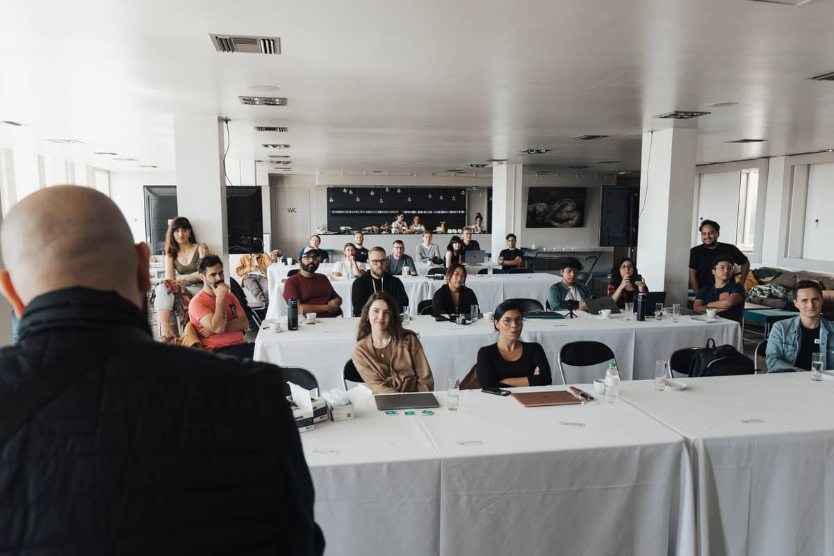 A group of adults seated at tables in a bright room, facing an instructor, suggesting a coffee workshop or class setting.
