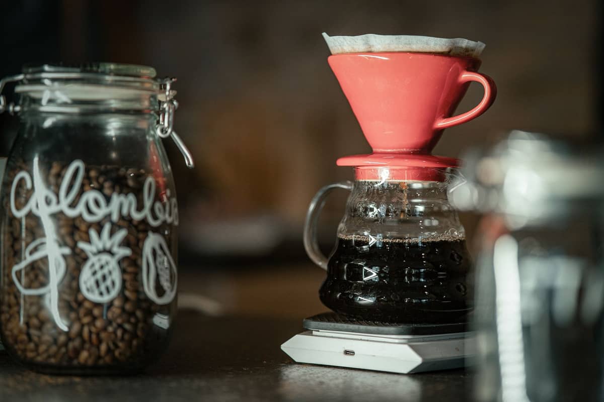 A jar labeled 'Colombia' filled with coffee beans sits beside a pour over coffee maker brewing fresh coffee on a counter.