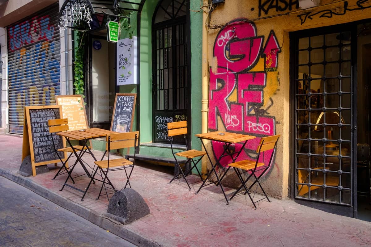 Outdoor seating area of a small urban coffee shop with graffiti and handwritten chalkboard, set in a colorful city street.
