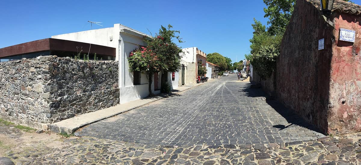 วิวตามถนน Calle de los Suspiros ใน Barrio Histórico ของ Colonia del Sacramento แสดงให้เห็นถนนปูหิน กำแพงหินประวัติศาสตร์ และบ้านสไตล์โคโลเนียลใต้ท้องฟ้าสีฟ้าใส
