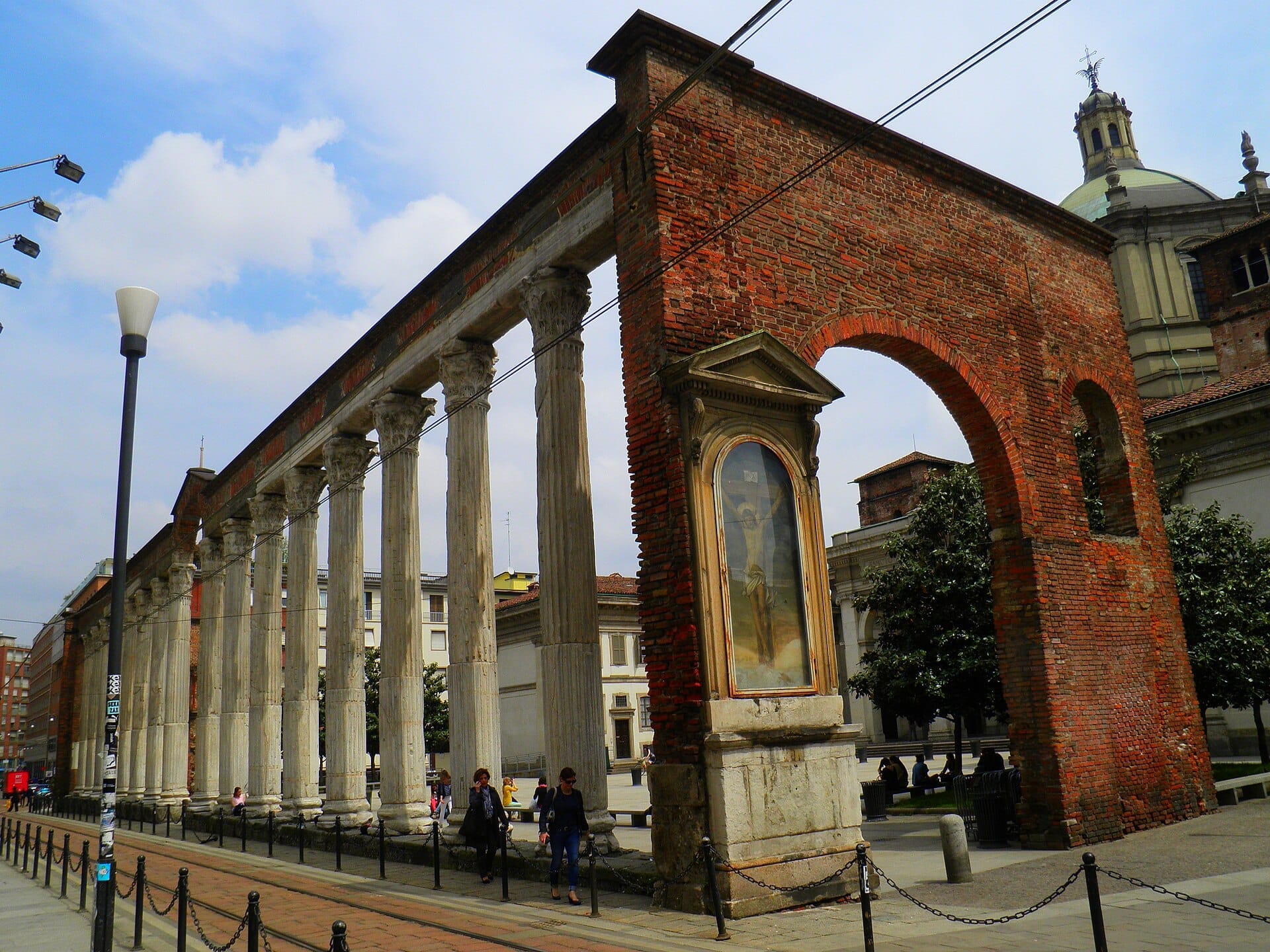 Le Colonne di San Lorenzo a Milano, con sedici antiche colonne corinzie in marmo e archi in mattoni rossi, sotto un cielo parzialmente nuvoloso.