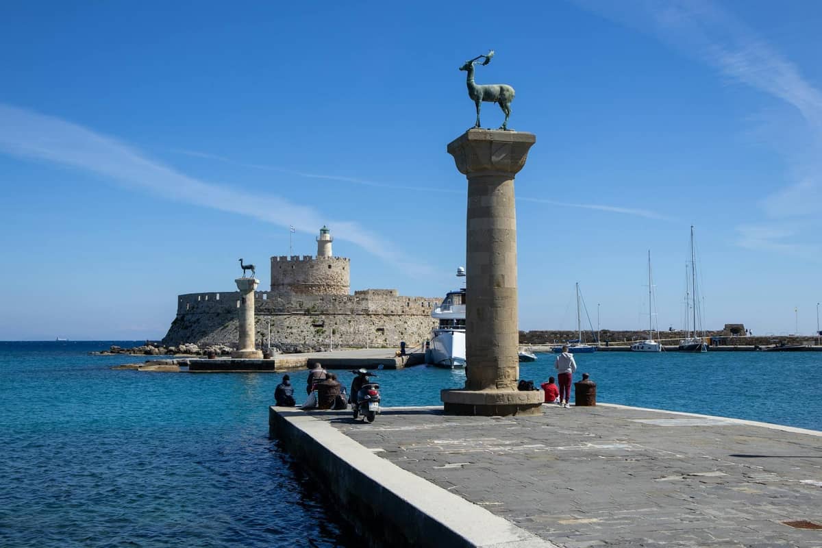 Mandraki Harbour in Rhodes with stone columns topped by deer statues and the medieval Fort of St. Nicholas in the background.