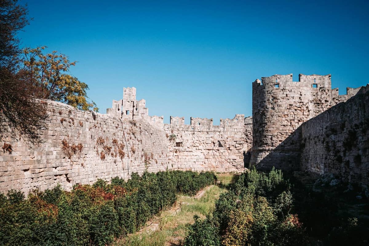 Medieval stone walls and towers surrounded by greenery and blue sky near Mandraki Harbour in Rhodes.