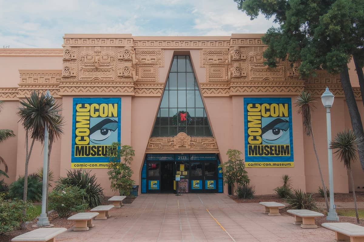 มุมมองด้านหน้าของ Comic-Con Museum ใน Balboa Park พร้อมด้วยด้านหน้าอาคารสีแทนที่ประณีต หน้าต่างบานใหญ่ ป้ายแบนเนอร์สะดุดตา ต้นปาล์ม และม้านั่งริมทางเดิน