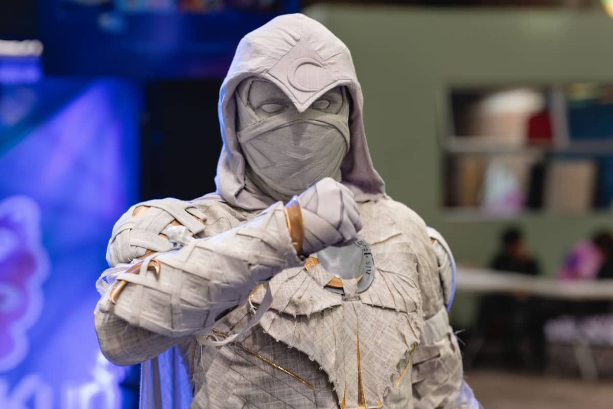 Ein Cosplayer im Kostüm eines maskierten Superhelden steht in einer Messehalle einer Comic-Convention, im Hintergrund sind bunte Messestände verschwommen zu sehen.