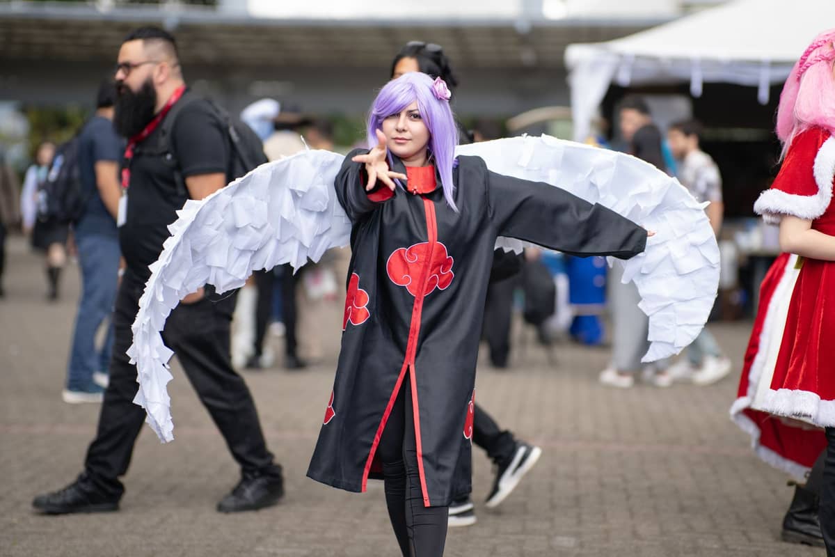 Cosplayer in einem aufwendigen Kostüm mit großen weißen Flügeln, der draußen unter anderen Comic-Con-Besuchern in Alltags- und Kostümkleidung steht.