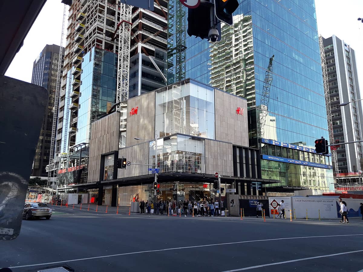 Blick von der Straße auf das Einkaufszentrum Commercial Bay im Auckland CBD mit dem H&M-Store und modernen Glasgebäuden bei Tageslicht.