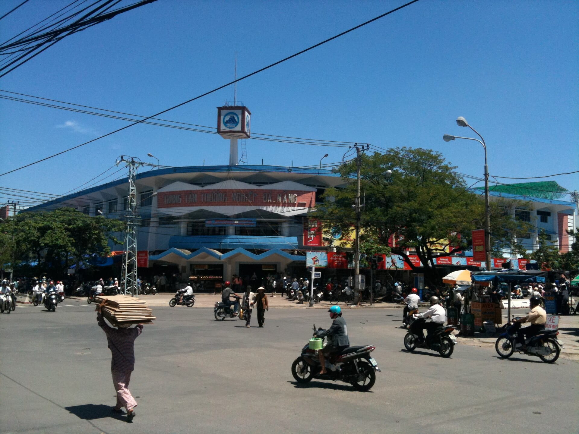 มุมมองภายนอกของตลาดก๊อน (Chợ Cồn) เมืองดานัง ยามแดดจ้า มีผู้คน มอเตอร์ไซค์ และต้นไม้ ใกล้ทางเข้าตลาดหลังคาน้ำเงินขาวขนาดใหญ่