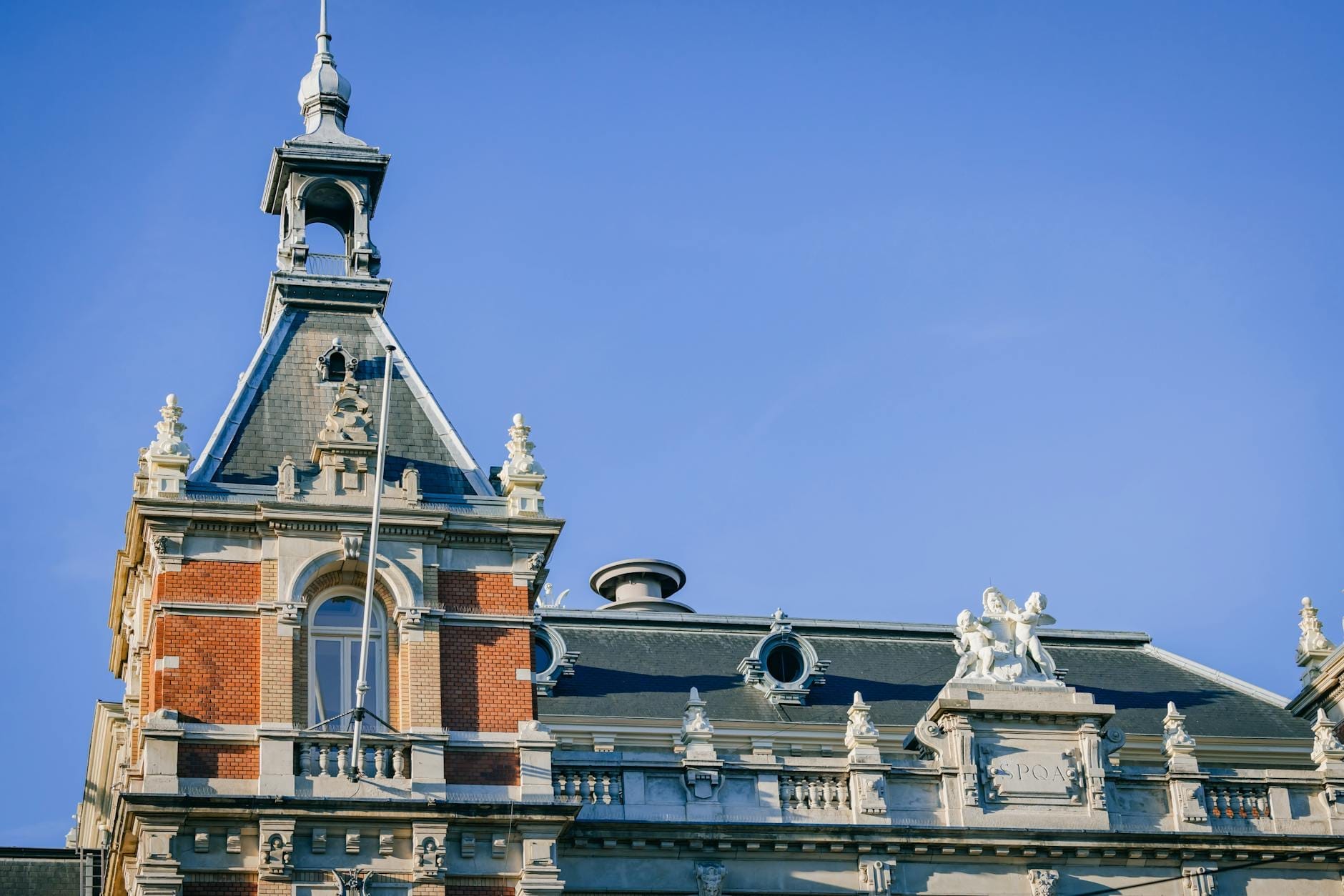 Sección superior ornamentada de un elegante edificio de sala de conciertos en Ámsterdam, con estatuas decorativas, arquitectura detallada y un cielo azul despejado de fondo.