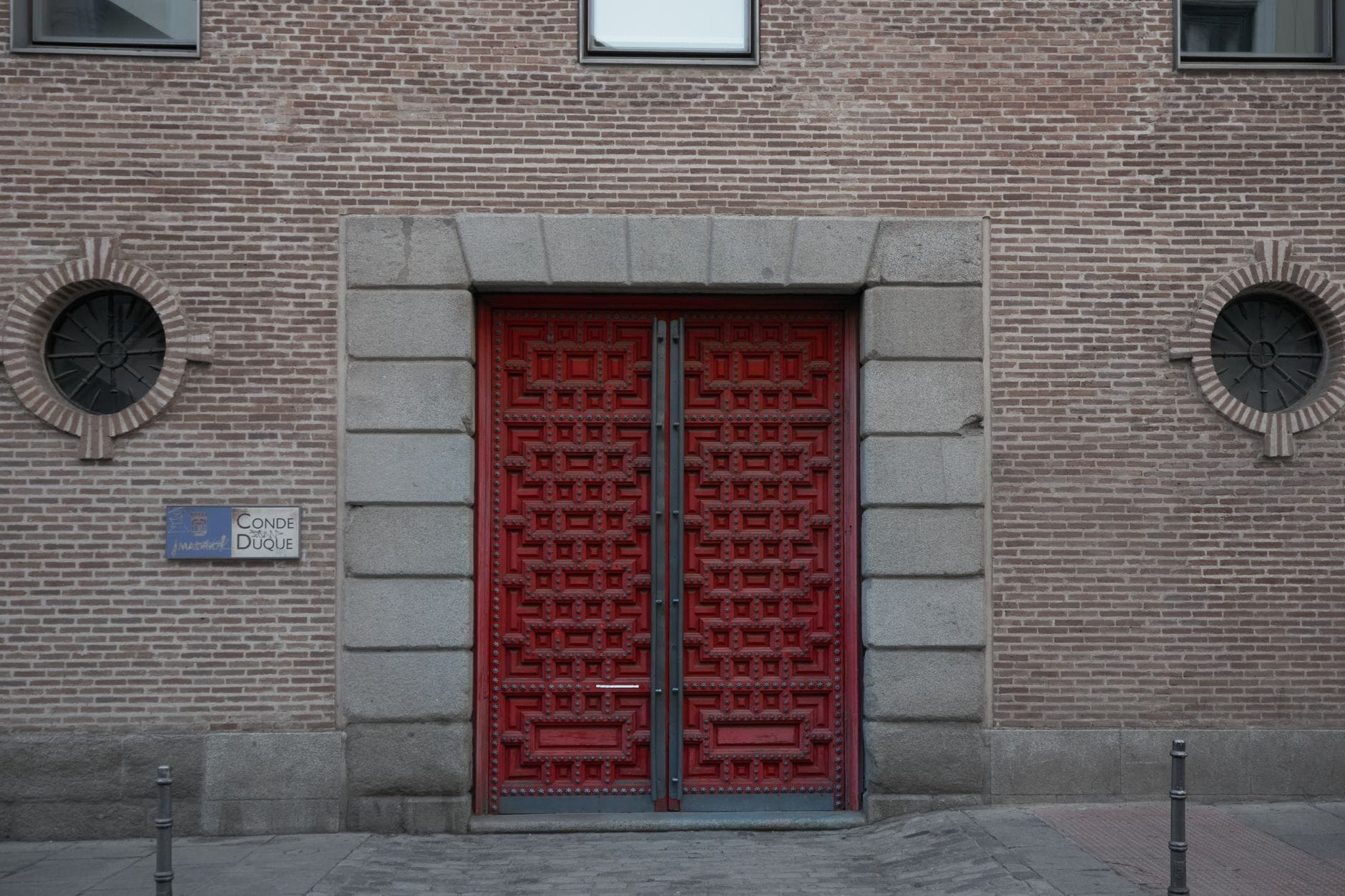 Grandi portoni ornamentali rossi incastonati nella facciata storica in mattoni con cornice in pietra e finestre circolari del Centro Cultural Conde Duque a Madrid.