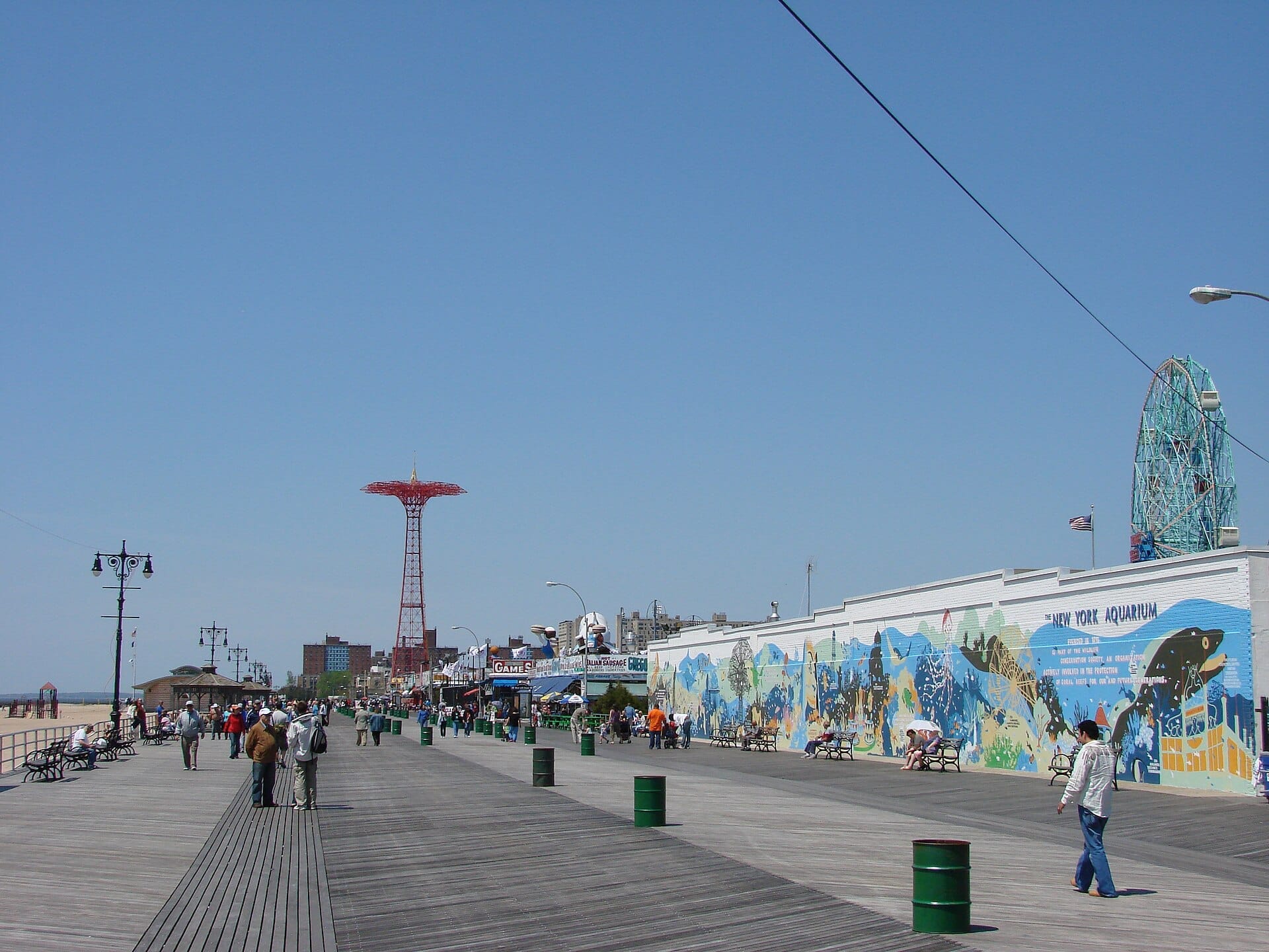 Visitatori che passeggiano lungo la Coney Island Boardwalk con cielo blu, un murale sulla parete dell'acquario, la torre Parachute Jump e la Wonder Wheel sullo sfondo.
