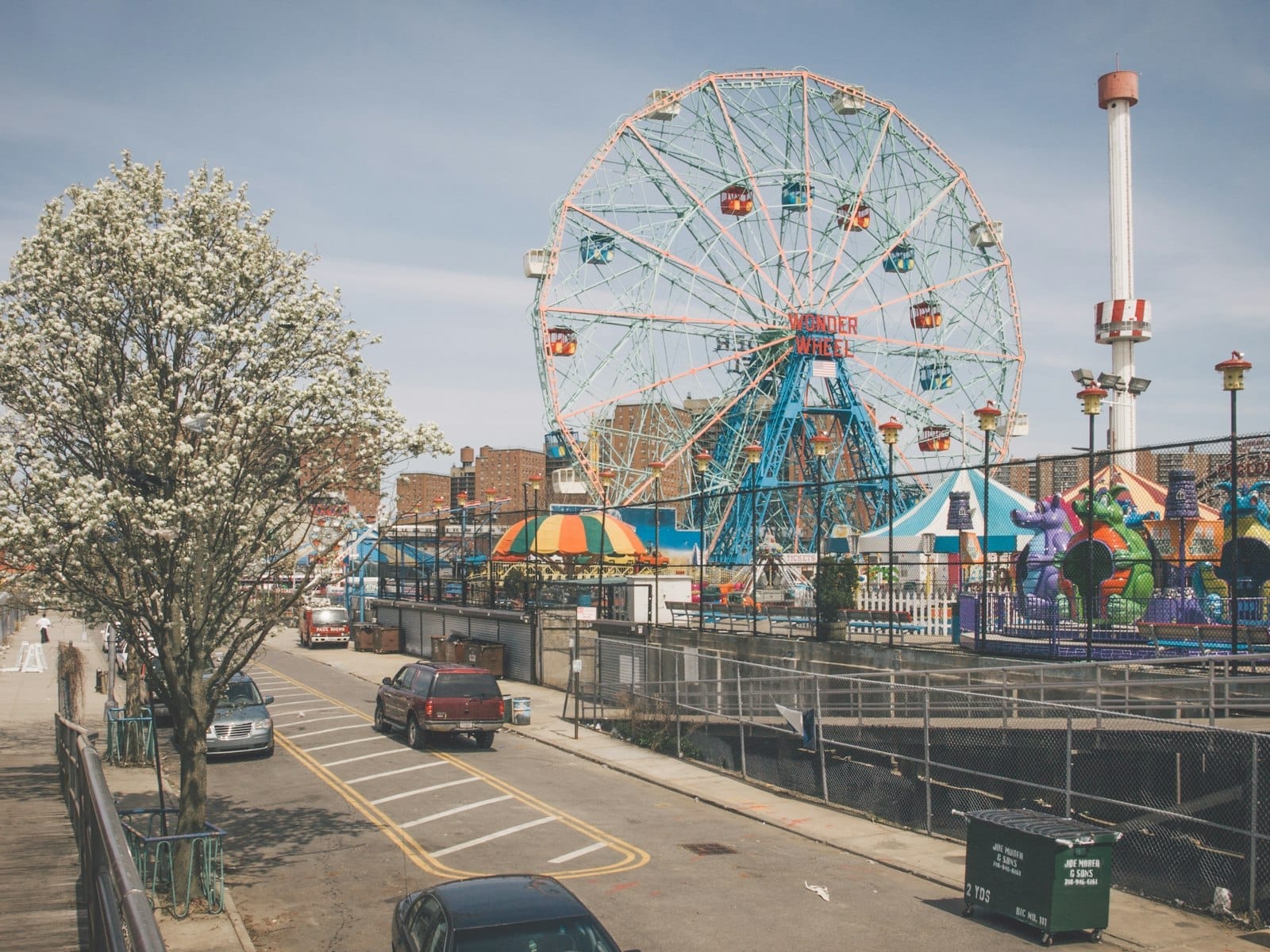 ภาพมุมกว้างสวนสนุกโคนีย์ไอแลนด์ เห็นชิงช้าสวรรค์ Wonder Wheel ต้นไม้ผลิบาน และเครื่องเล่นสีสันสดใสในวันที่แดดจ้า