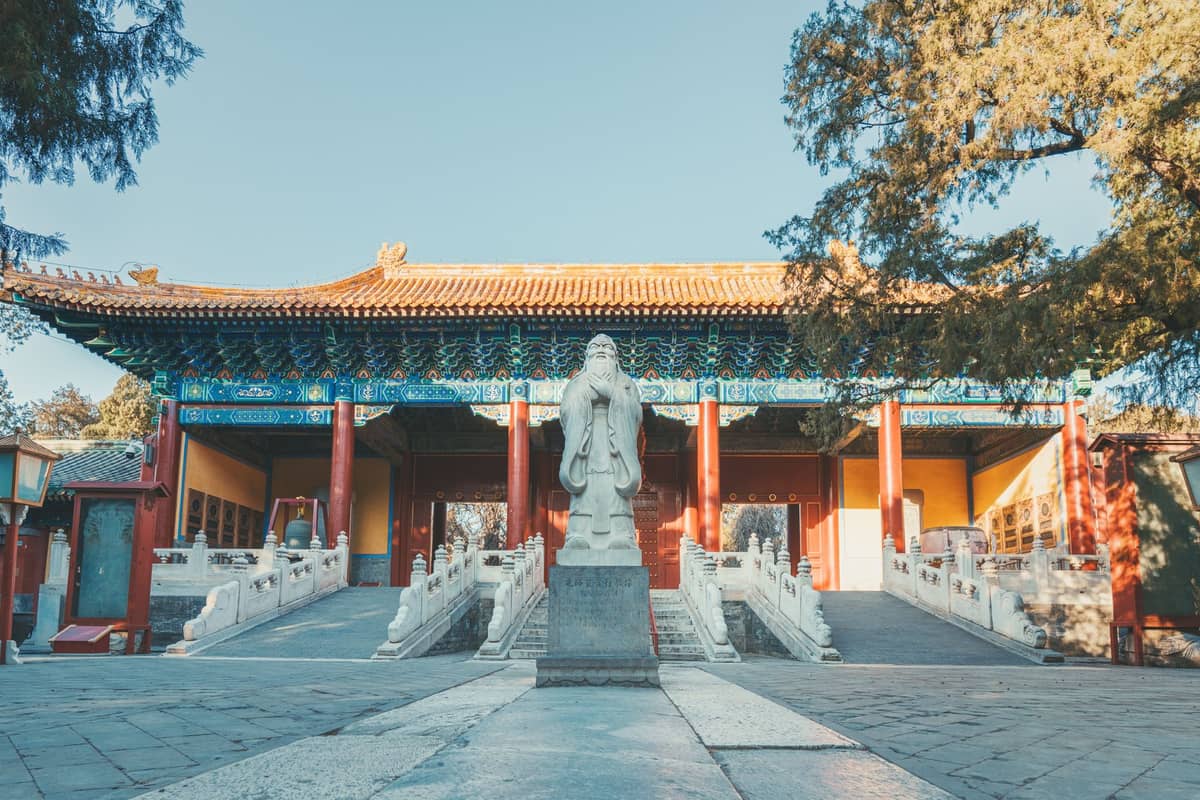 Weitwinkelbild des Eingangs zum Konfuzius-Tempel in Peking mit einer markanten weißen Konfuzius-Statue, einem verzierten Tor, roten Säulen und schattigen Innenhöfen.